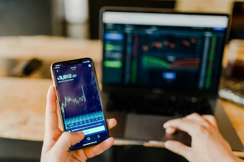 Investor checking a stock trading app on a smartphone next to live market charts on a laptop