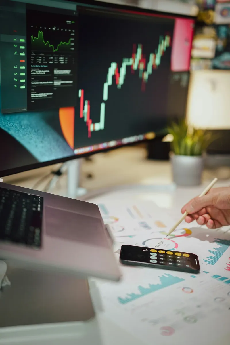 Computer monitor showing a bullish candlestick chart beside printed financial reports and a calculator